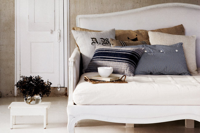 A white daybed with striped and monogrammed pillows beside a small white door, soft and serene